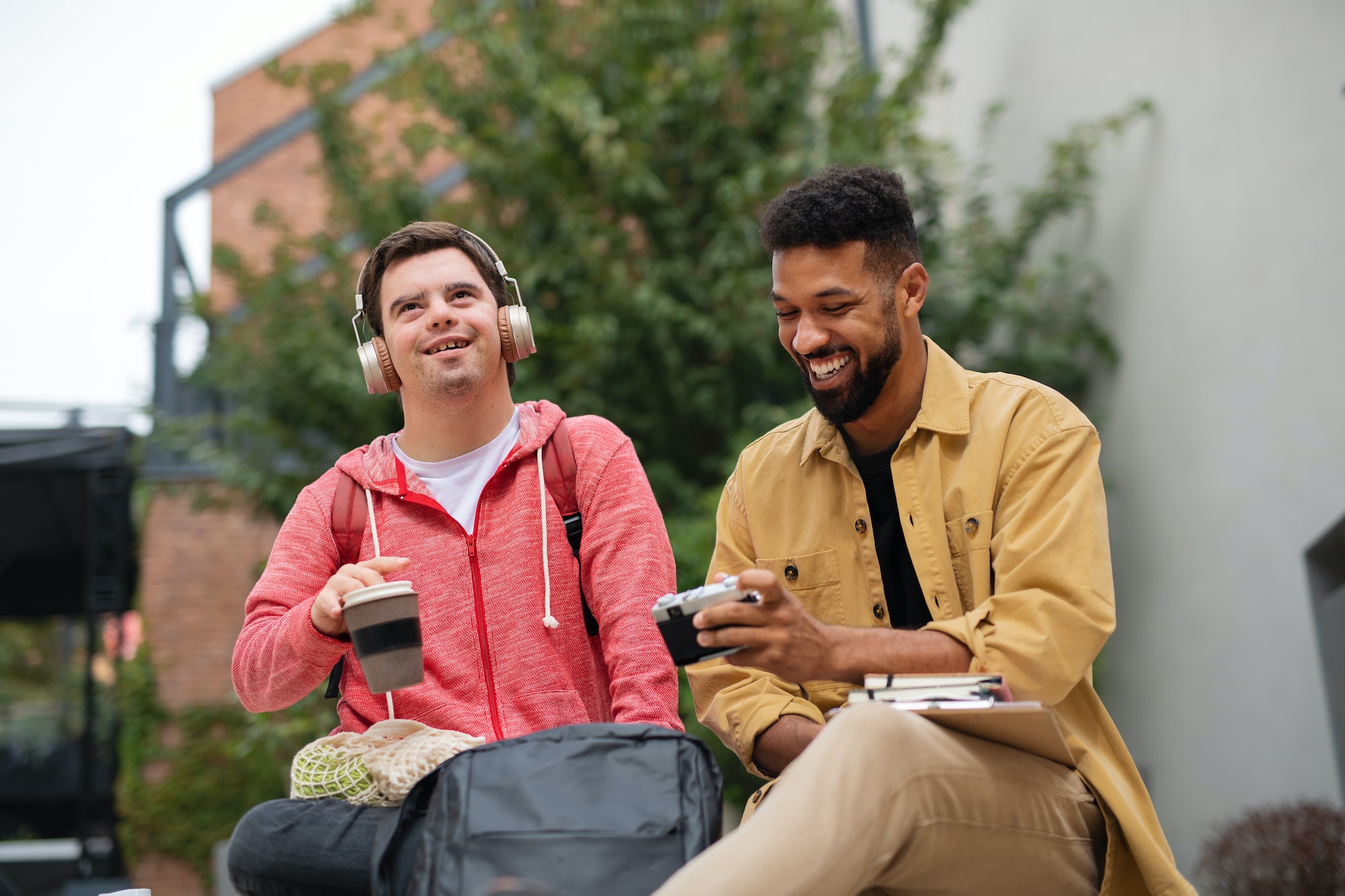 Young man student with Down syndrome and his mentoring friend sitting outdoors in campus area